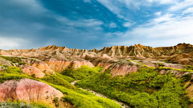 A vibrant landscape featuring steep, eroded rock formations (badlands) covered in bright green vegetation under a blue sky with white clouds.