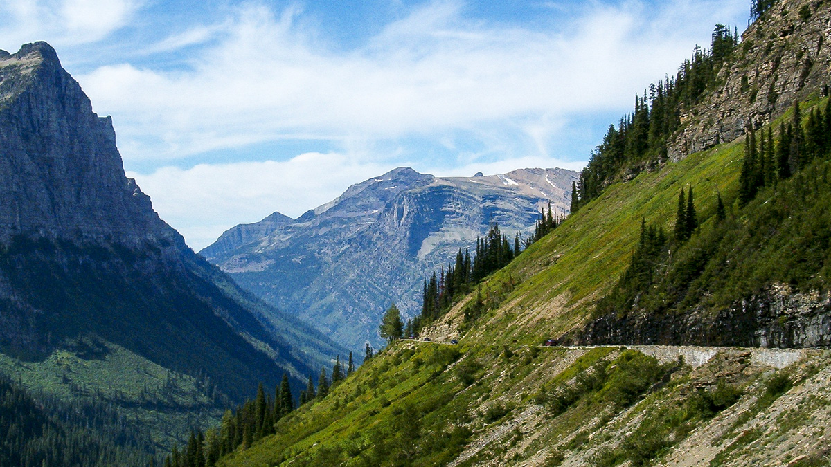 A scenic mountain landscape featuring steep, green, tree-covered slopes leading up to rugged, snow-capped peaks under a blue sky with white clouds. A winding road is visible on the lower right slope.