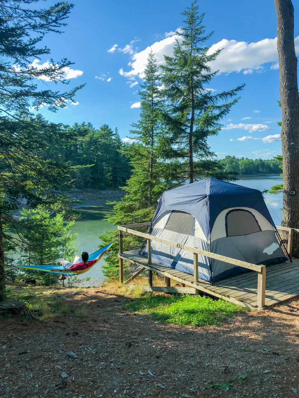 A tent is pitched on a wooden deck next to a calm lake at Acadia National Park. A a hammock is slung between two trees nearby, with a man sitting in it.
