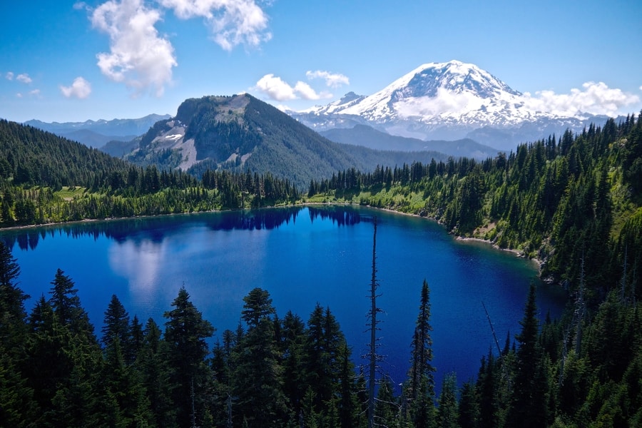 Mount Rainier National Park A vibrant blue lake surrounded by evergreen trees and mountains, with the snow-caped Mount Rainier visible in the background under a partly cloudy sky.