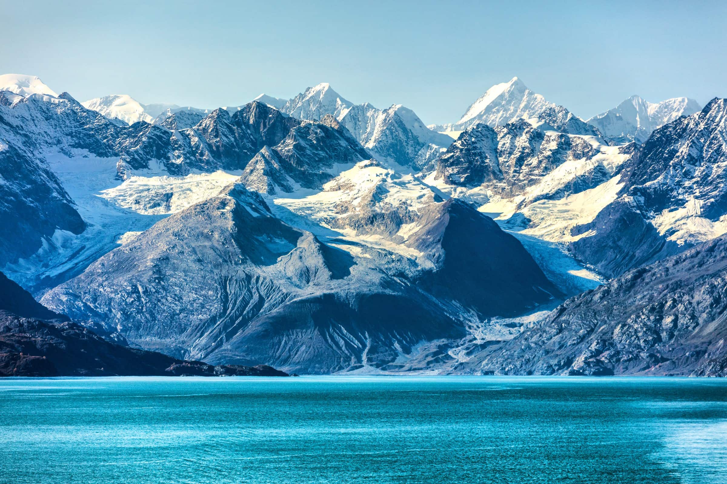 A scenic view of large, snow-capped mountains and glaciers overlooking a body of aqua-colored water in Glacier Bay.