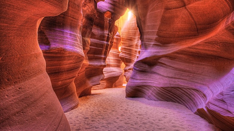 A vibrant photograph of the inside of Antelope Canyon, featuring wavy sandstone formations in shades of orange, red, pink, and purple, with sunlight streaming in from above.