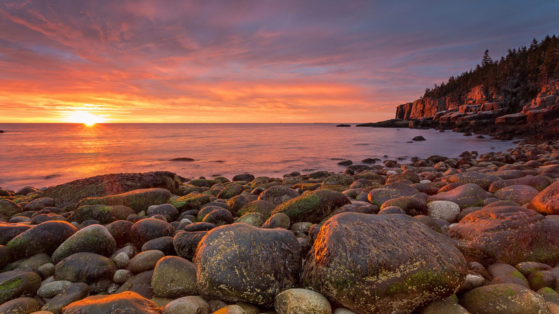 Sunrise over a rocky coastline with large stones in the foreground.