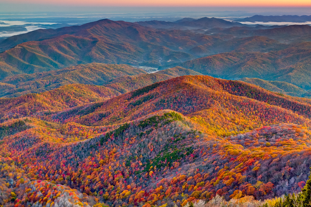 An aerial view of the Great Smoky Mountains National Park during autumn, showing rolling hills covered in vibrant orange, yellow, and green foliage under a misty sky.
